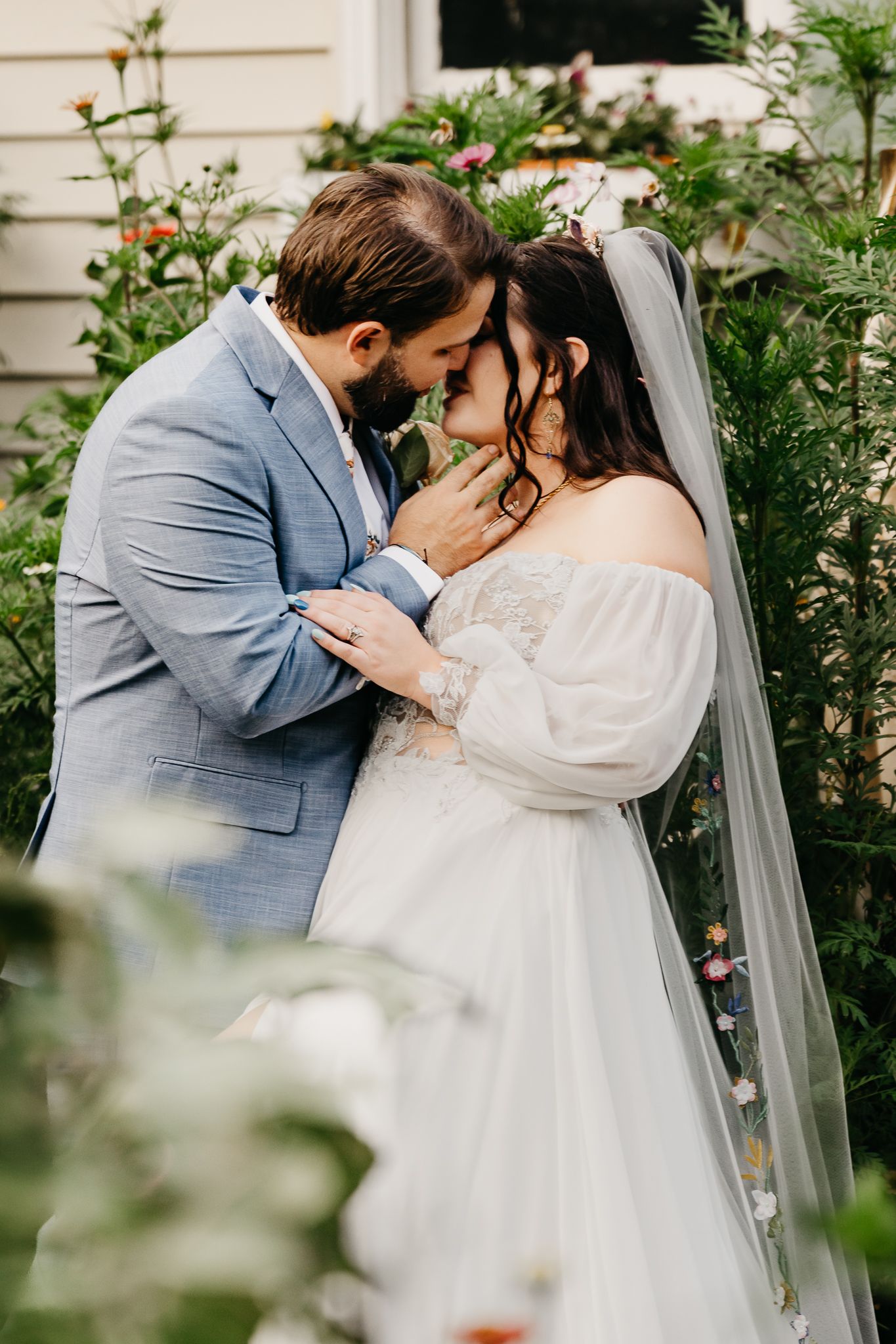 Romantic wedding couple shares intimate kisses in garden setting wearing gray suit and white dress with flowing veil.