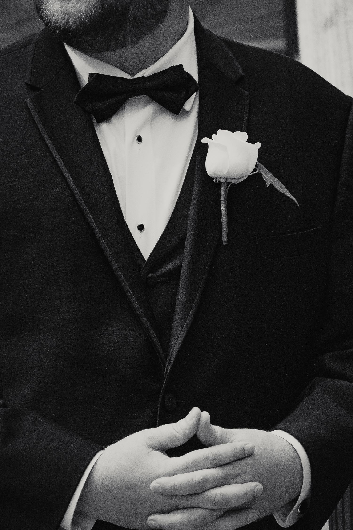 Black and white photo of a groom in a tuxedo with bow tie and white rose boutonniere, hands clasped.