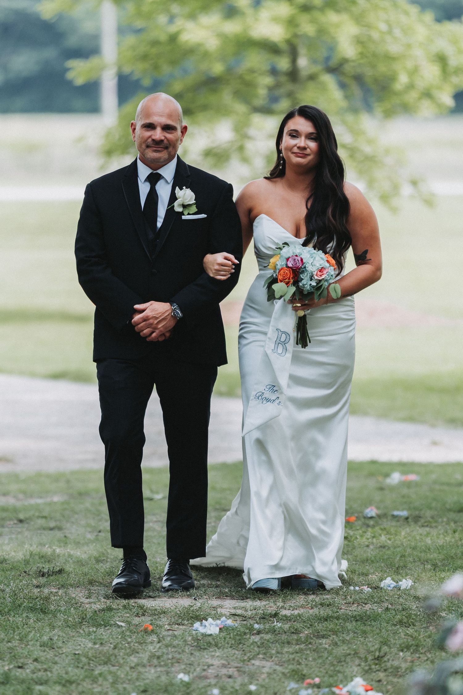 Bride in white strapless gown holding colorful bouquet walks down aisle with man in black tuxedo at outdoor wedding.