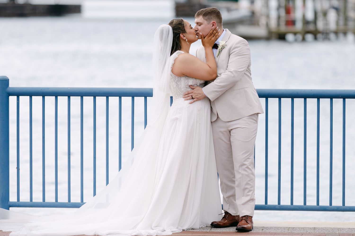 A bride and groom share a romantic kiss on a waterfront dock, with blue railings and calm water in the background.