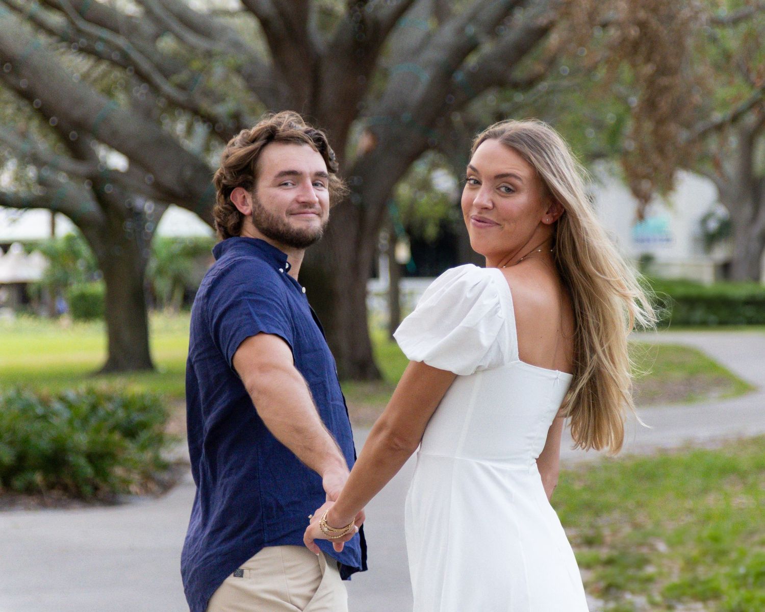 Romantic couple holding hands and looking back while walking through a tree-lined park.