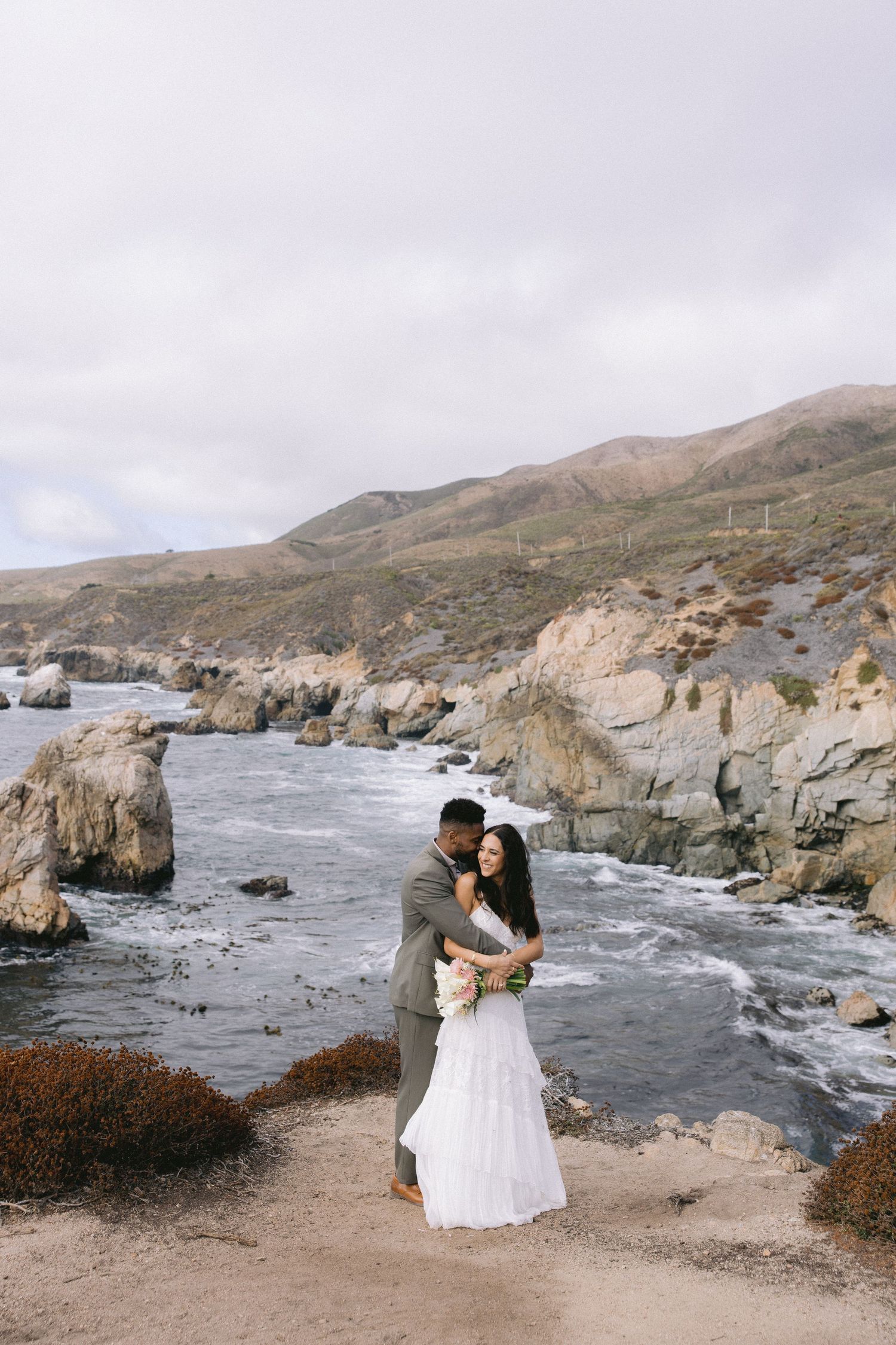 A couple in wedding attire embrace on a cliff overlooking the rocky coastline at Big Sur, California.