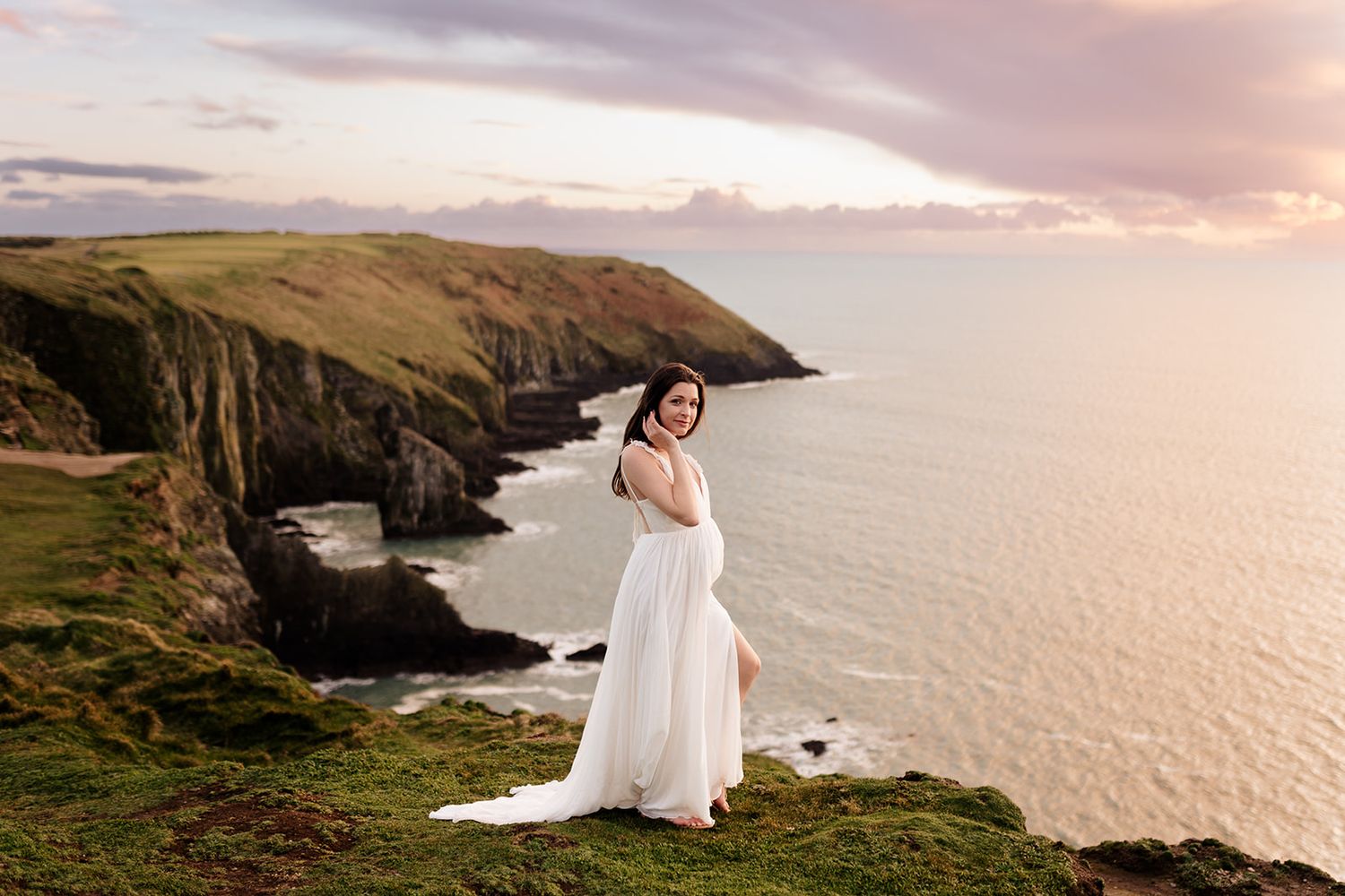 Bride in flowing dress poses on dramatic coastal cliffs at sunset.