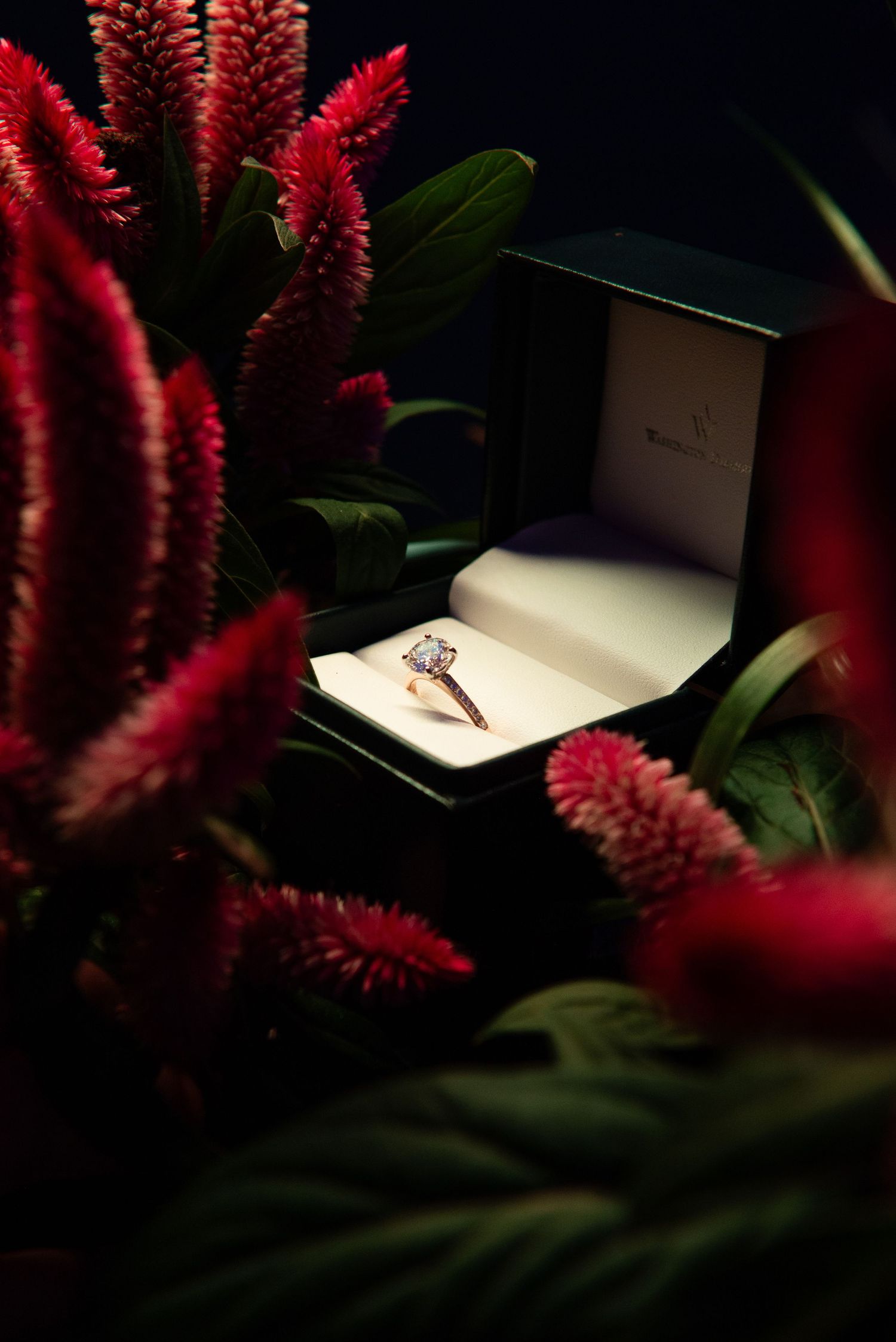 Diamond engagement ring in black box surrounded by red flowers and greenery.