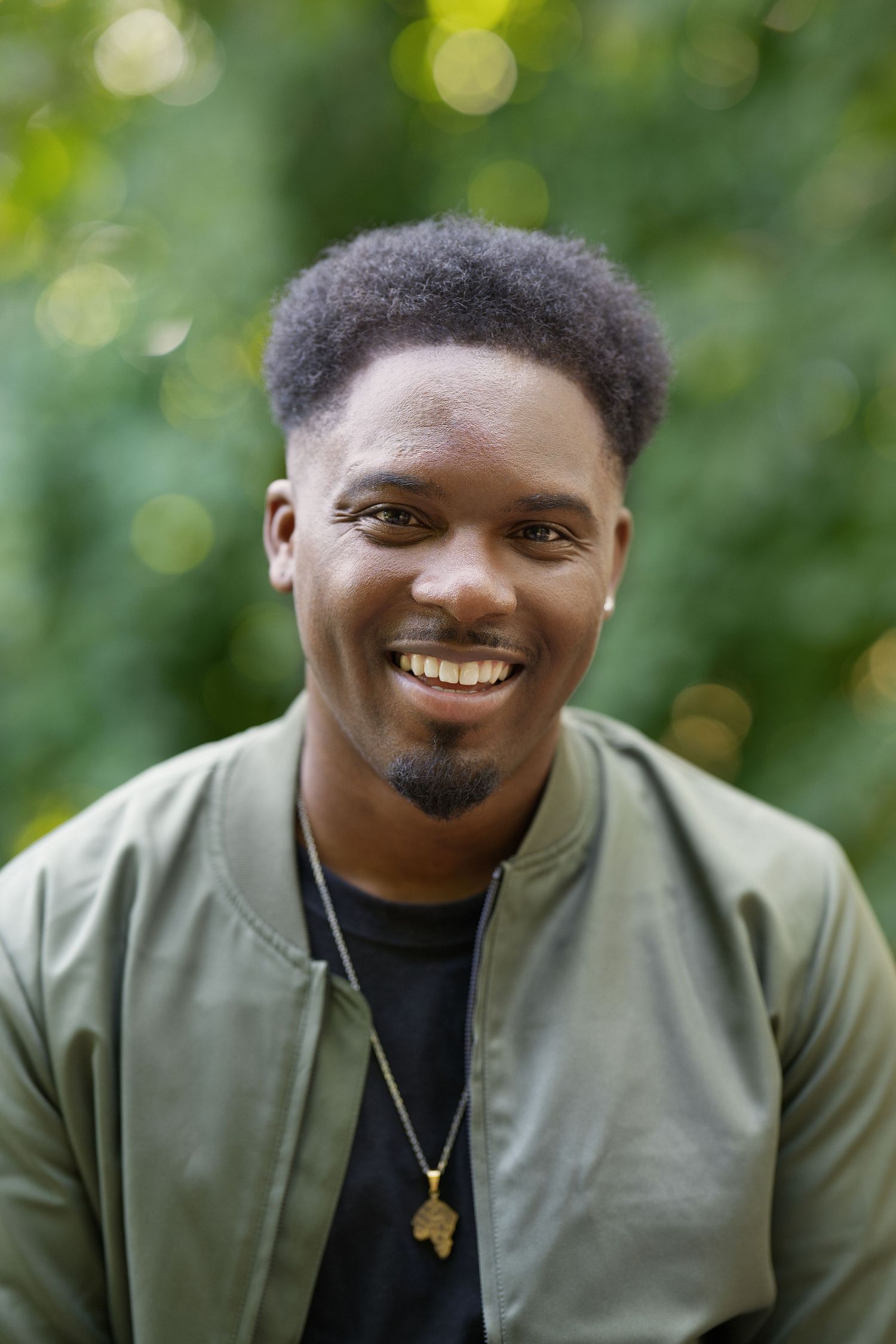 Outdoor portrait of smiling person in olive jacket with green bokeh background.