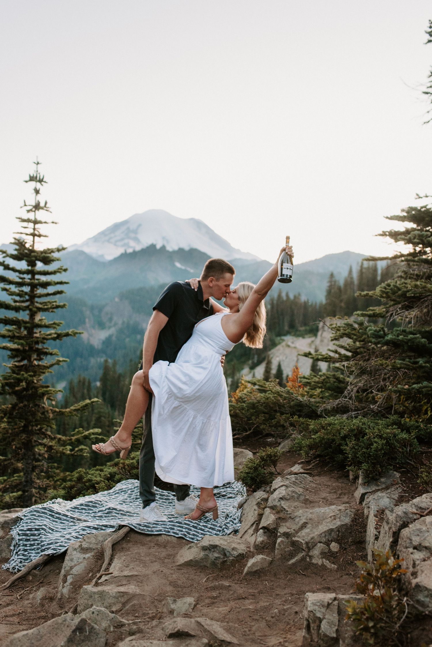 Couple embracing on rocky mountain overlook with evergreen trees and scenic peaks in background.