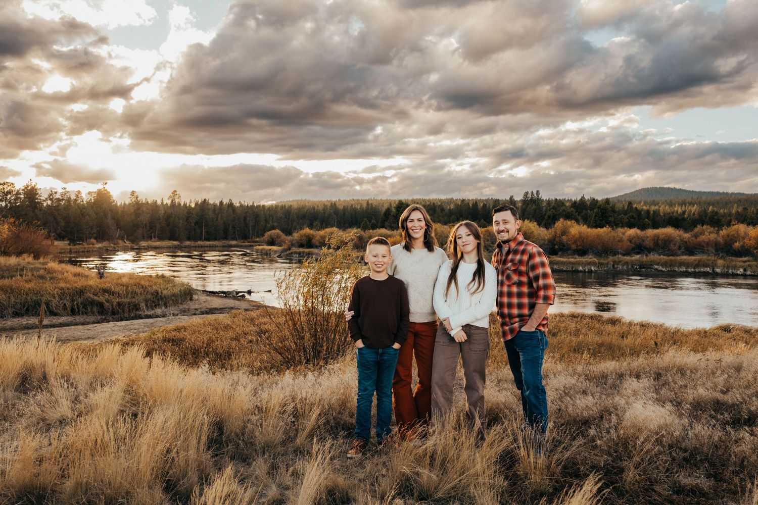 A family stands on a grassy hill overlooking a serene river landscape at sunset with dramatic cloudy skies.