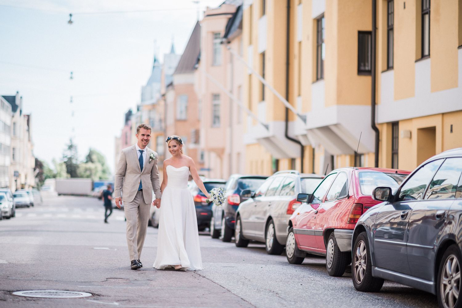 A newlywed couple walks down a European street lined with parked cars on a sunny afternoon.
