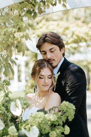 A romantic outdoor wedding moment with a couple embracing under green foliage and soft sunlight.