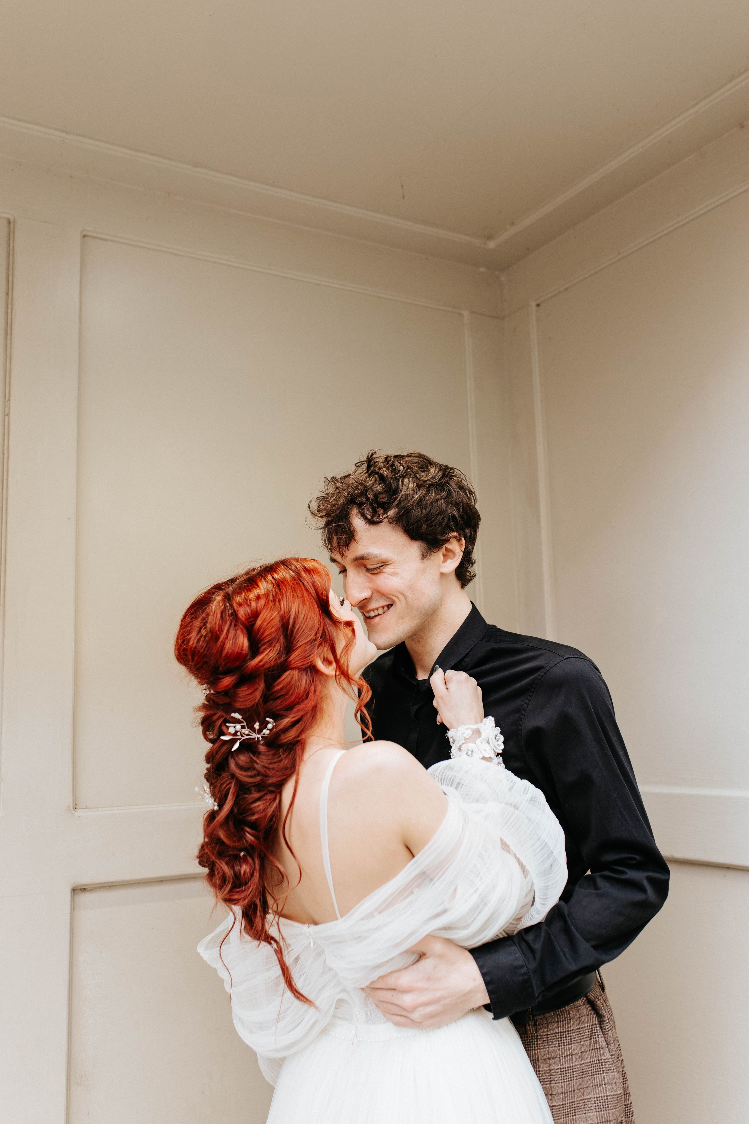 A romantic couple embraces against a white wall, with the woman wearing a white dress and having vibrant red hair.