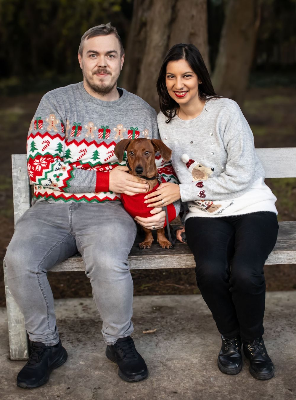 Couple in holiday sweaters sits on bench with a dog, wearing festive outfits for Christmas photo.