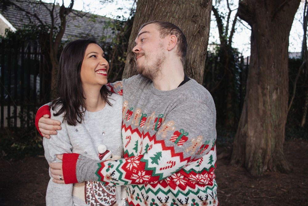 A couple wearing matching winter sweaters share a tender moment in front of trees during the holiday season.