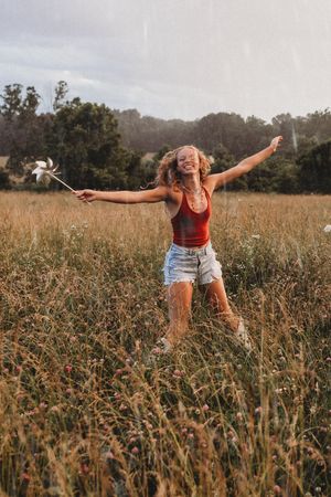 senior girl dancing in rain in the field with windmill jean shorts red tank top curly hair