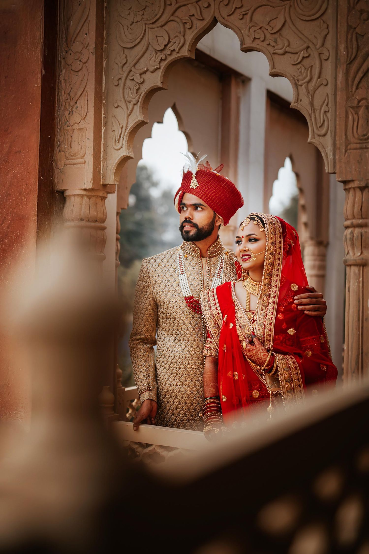 A couple in traditional Indian wedding attire pose beneath an ornate archway during a traditional ceremony.