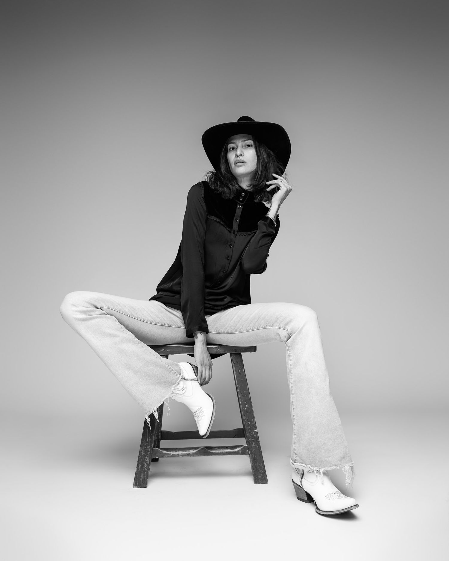 Artistic black and white portrait of person in hat and bell bottoms seated on stool.