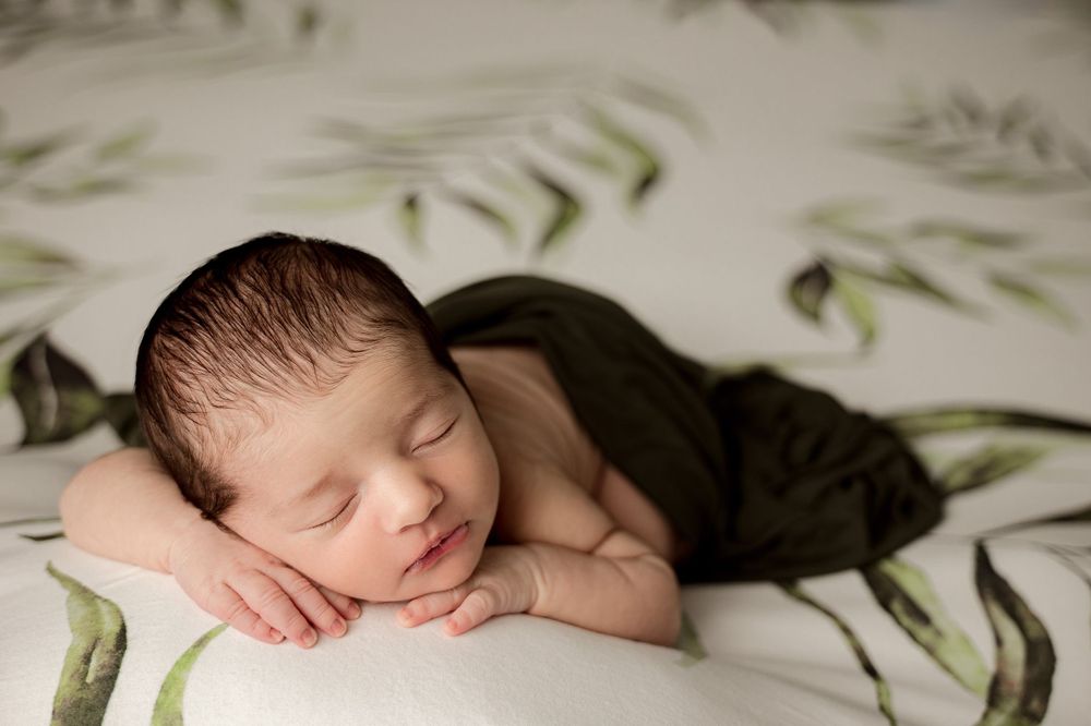 A sleeping newborn baby lies peacefully on a white surface with green leaf pattern background.