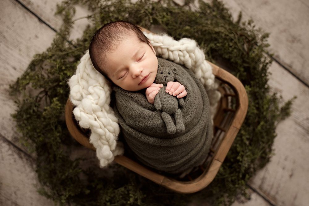 Sleeping newborn wrapped in gray swaddle rests in wooden basket surrounded by moss and white blanket.
