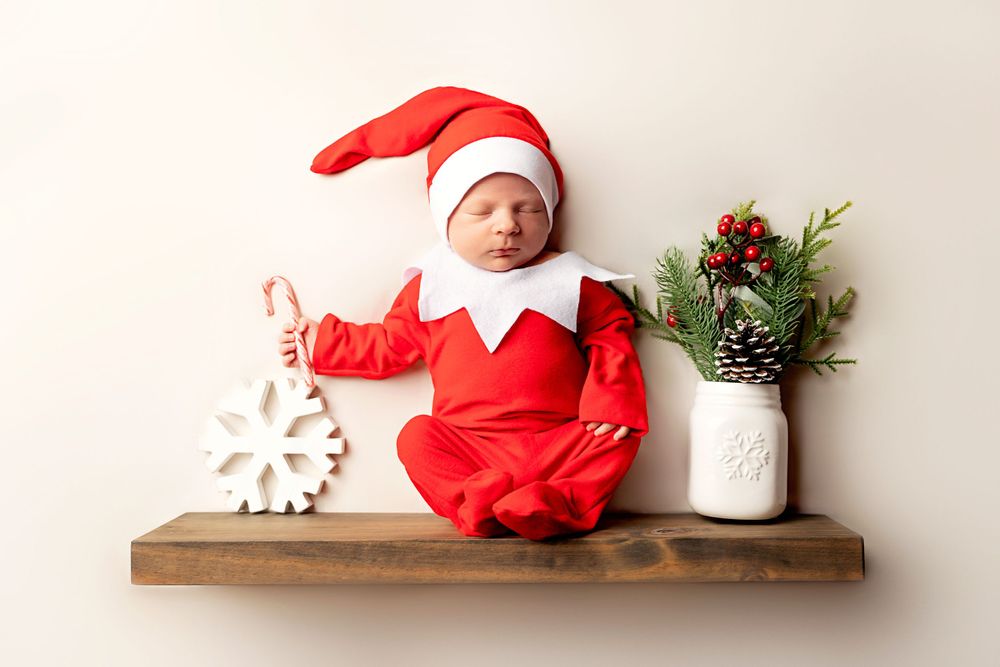 A festive holiday photo with a small figure in red elf costume sitting on wooden shelf with snowflake and pine decor.