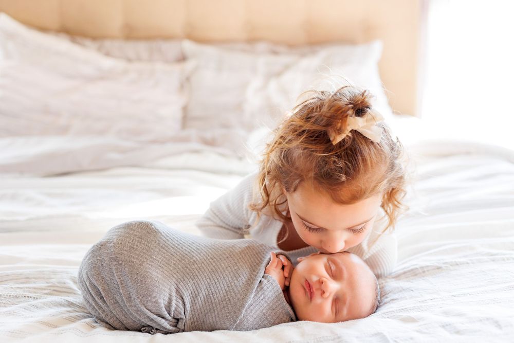 Tender sibling moment on white bedding with soft natural lighting.