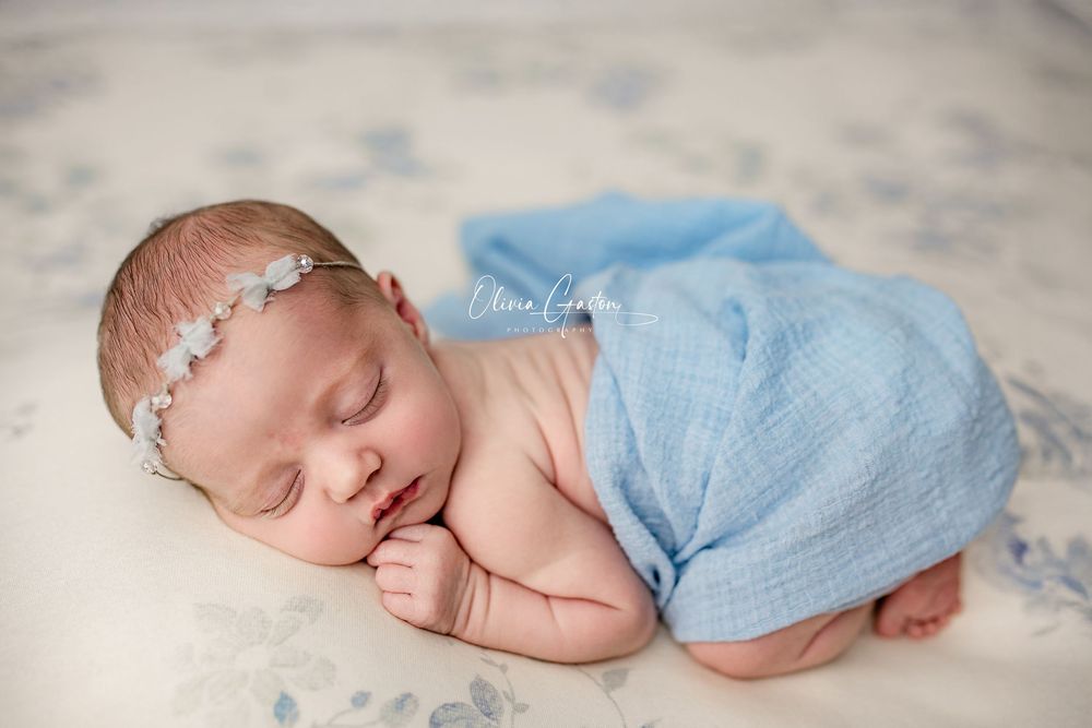 Sleeping newborn baby girl wrapped in soft blue fabric with a delicate floral headband on a light floral backdrop.