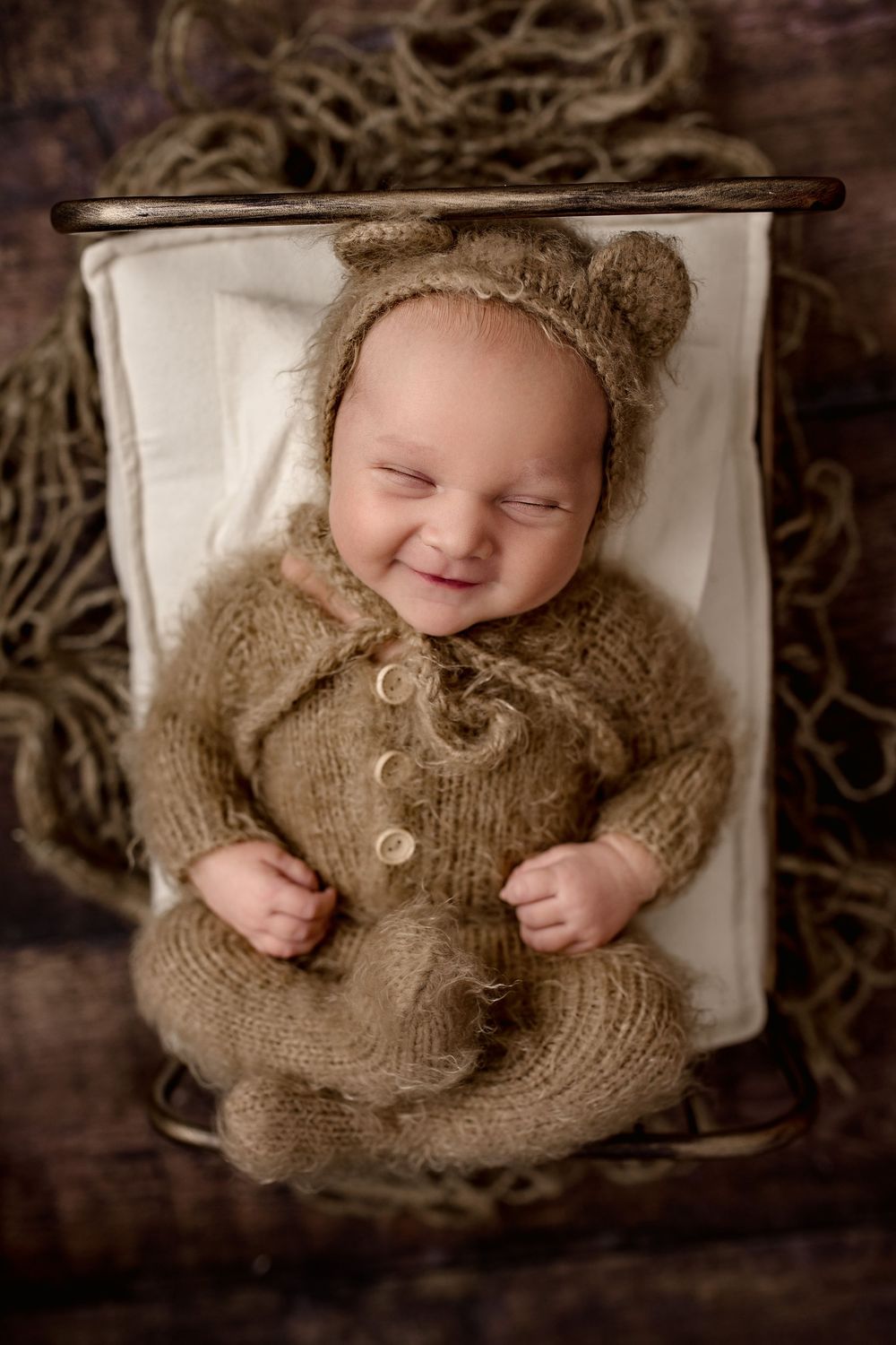 Sleeping baby wearing cozy knit brown bear outfit with ears lies on white cushion in rustic setting.