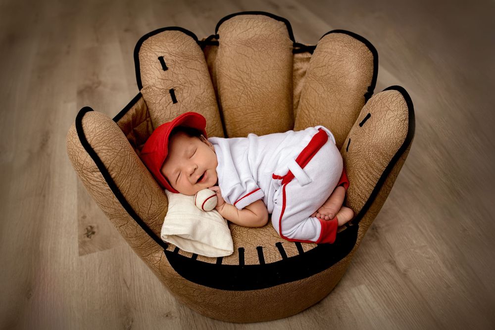 Sleeping baby wearing red and white outfit nestled in brown wooden bowl against neutral backdrop.
