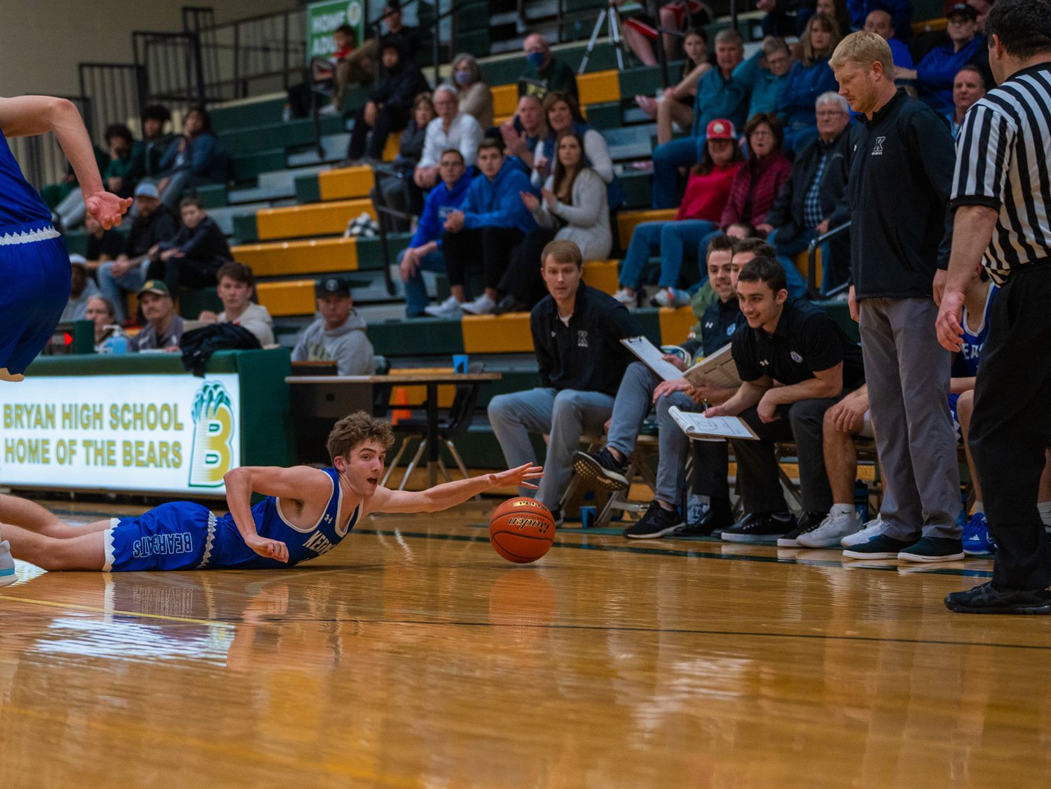 Player dives for loose basketball on gymnasium floor during competitive game with bench visible.
