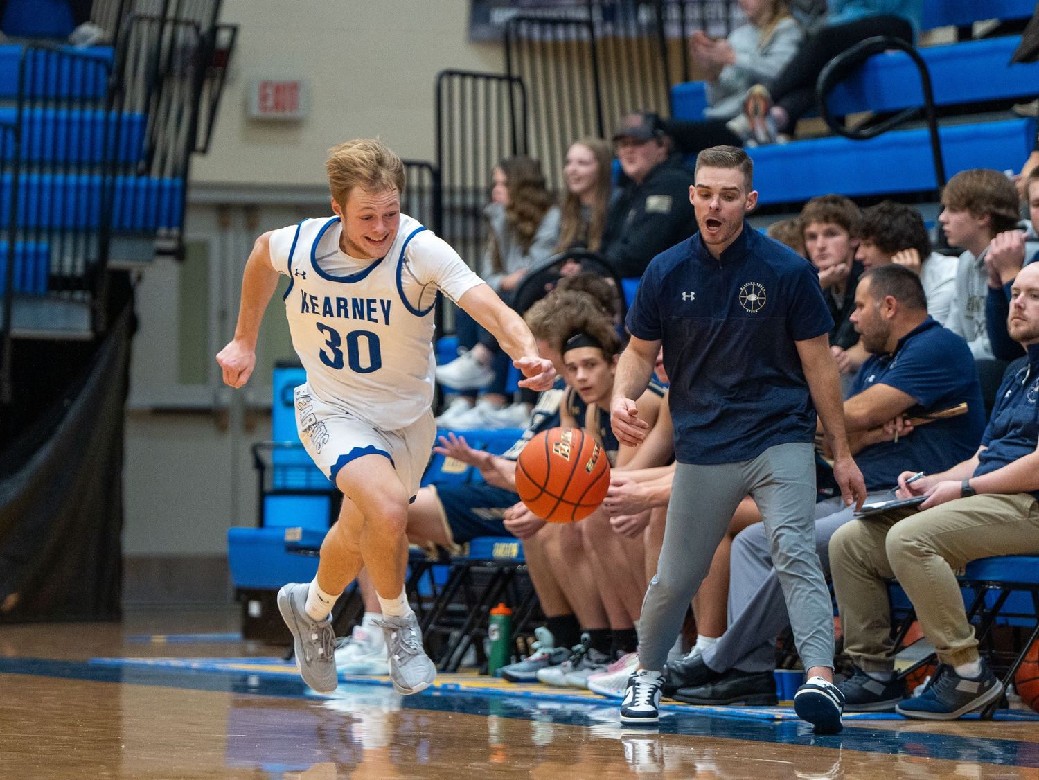 Basketball player dribbles along sideline while coach looks on during game action.