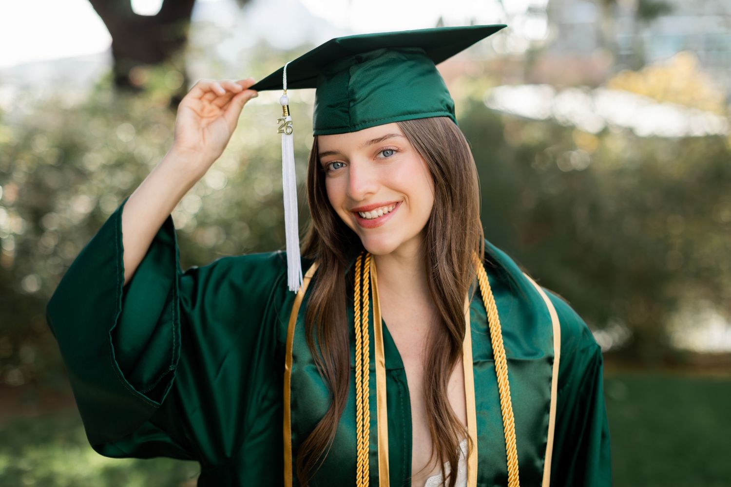 Person with long dark hair holding a green 2022 graduation sash outdoors in a sunlit field.