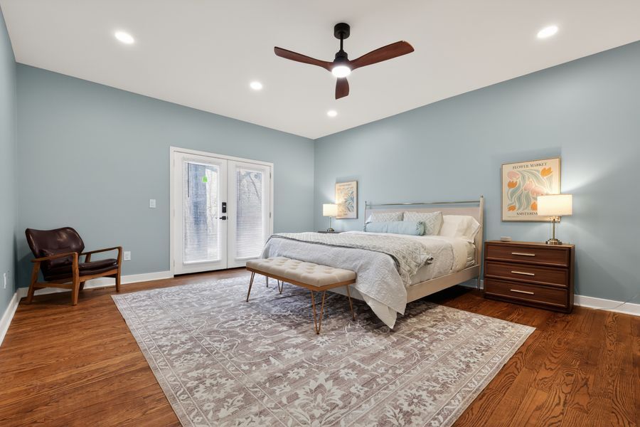 Spacious bedroom with light blue walls, hardwood floors, ceiling fan, French doors, and mid-century modern furniture on a patterned rug.