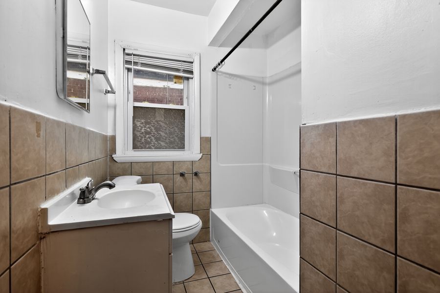 Small bathroom with brown tile surround bathtub, pedestal sink, toilet, and window providing natural light.