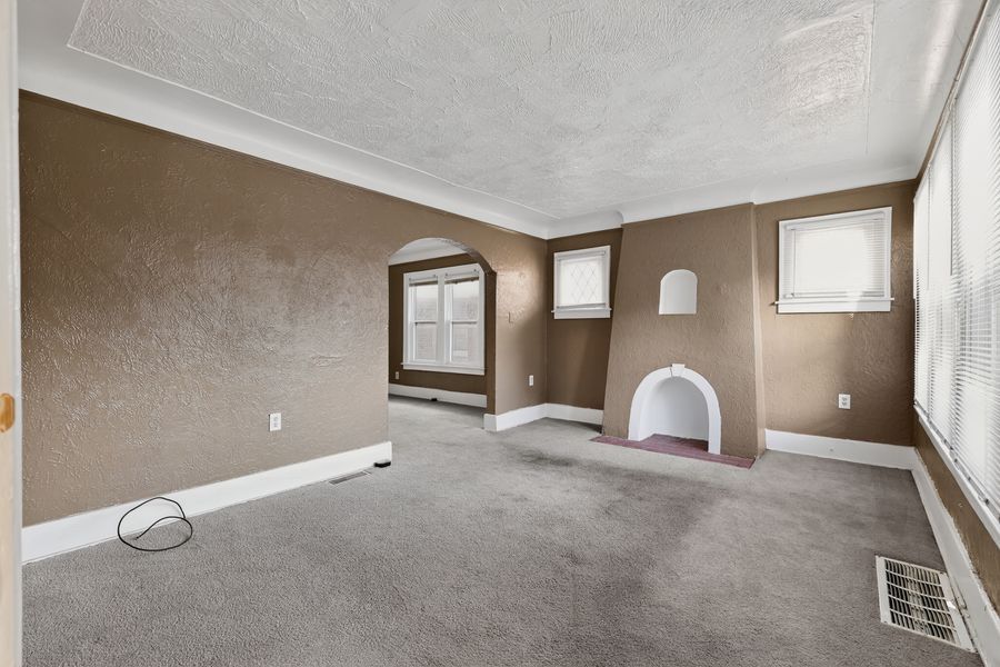 Empty living room with gray carpet, decorative fireplace, brown walls, crown molding, and two windows with shutters.
