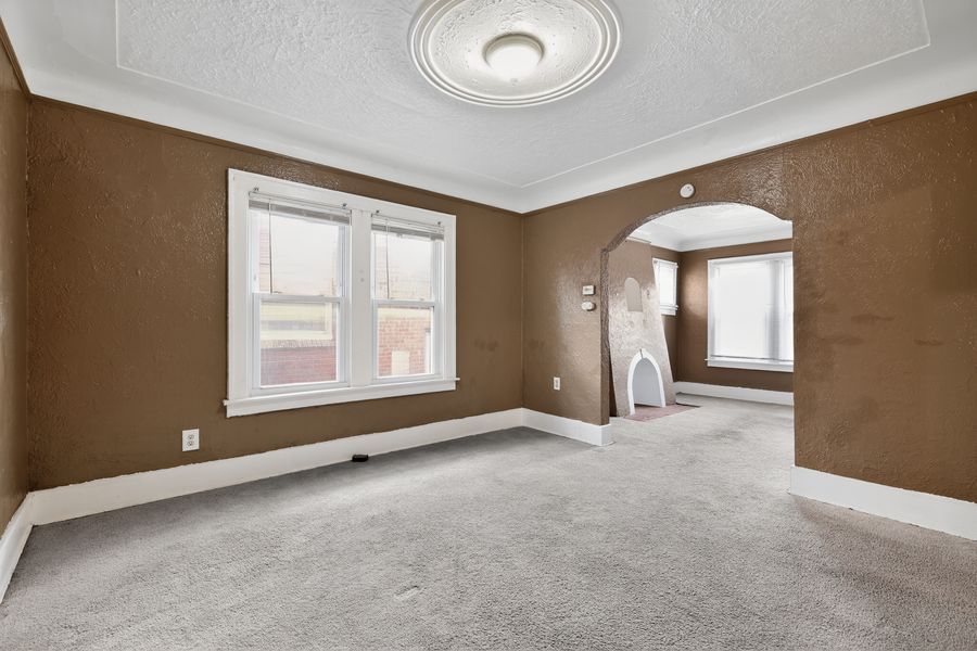 Brown-walled living room with gray carpet, crown molding, arched doorway, and two windows letting in natural light.
