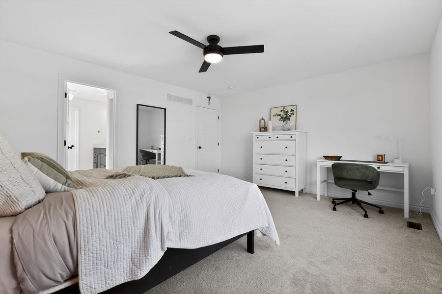 Bright bedroom with ceiling fan, plush carpet, white dresser, desk, and en-suite bathroom door in a modern home.