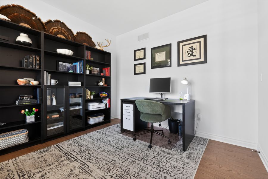 Home office with dark bookshelf, wooden desk, green chair, and framed wall art on hardwood floors.