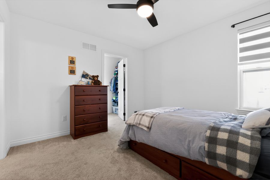 Cozy bedroom with gray plaid bedding, ceiling fan, dresser, and walk-in closet on carpet.