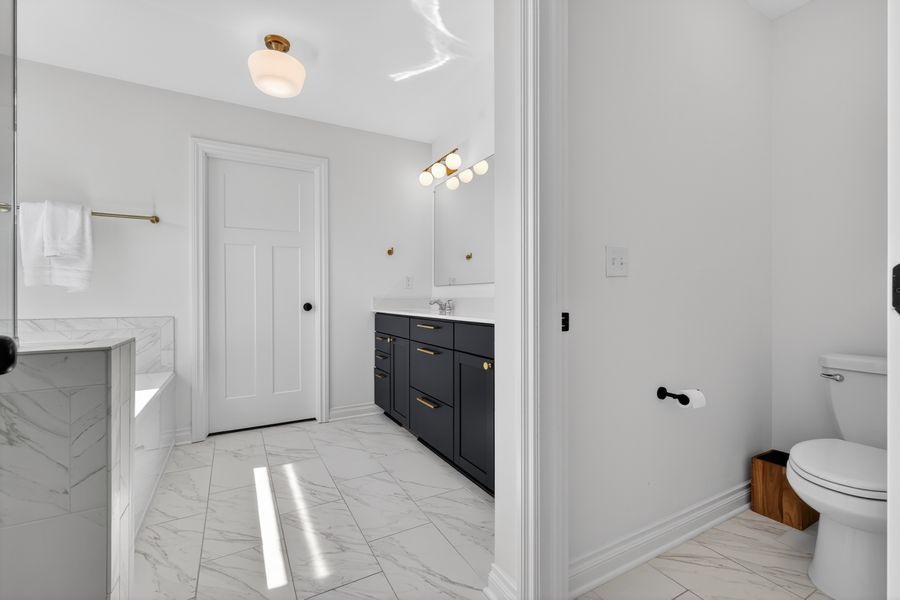 Modern white bathroom with marble tile floors, dark vanity, gold fixtures, and toilet.