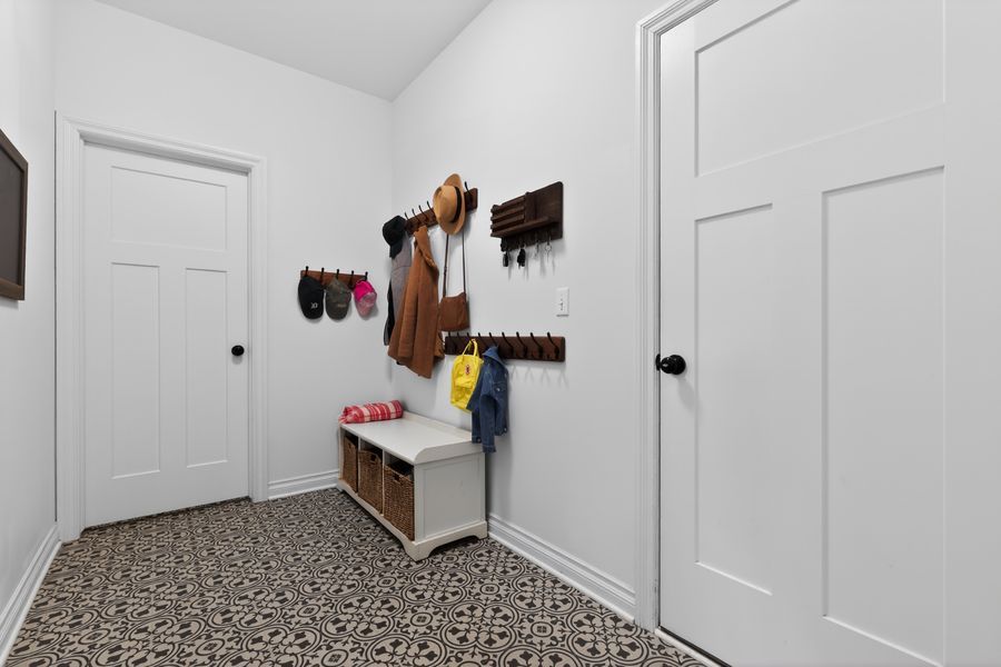 Mudroom with patterned tile floor, white walls, wall-mounted coat hooks with bags, and a small white storage bench.