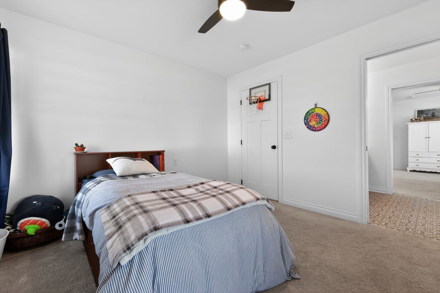 Cozy carpeted bedroom with a plaid twin bed, ceiling fan, basketball hoop decor, and white walls.