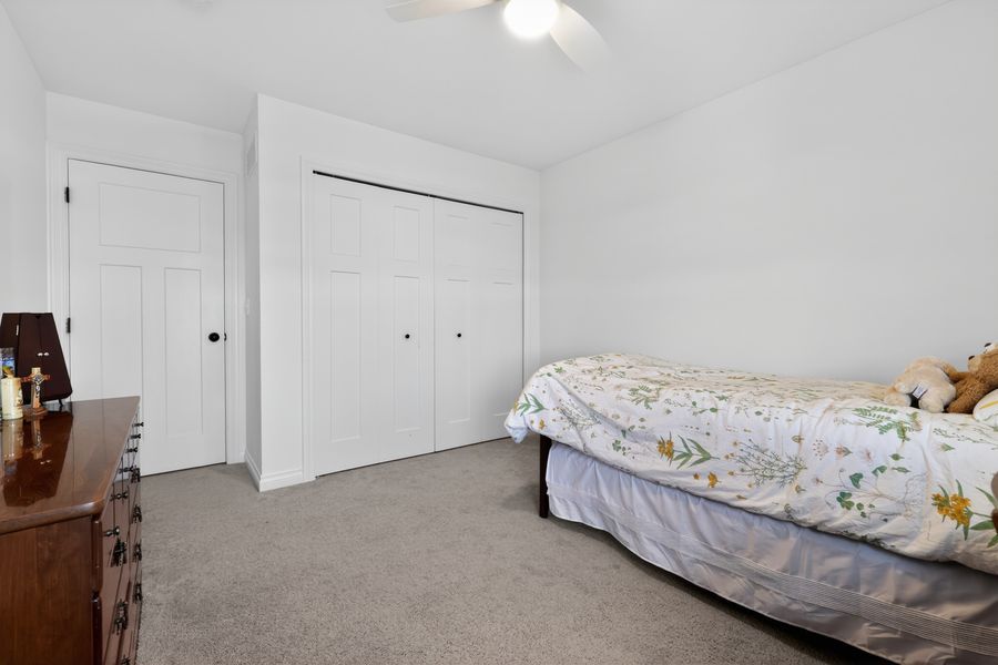 Cozy bedroom with floral bedding, beige carpet, white closet doors, ceiling fan, and a wooden dresser.