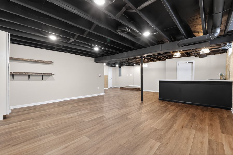 Finished basement with hardwood floors, exposed black ceiling, recessed lighting, white walls, and a dark kitchen bar area.