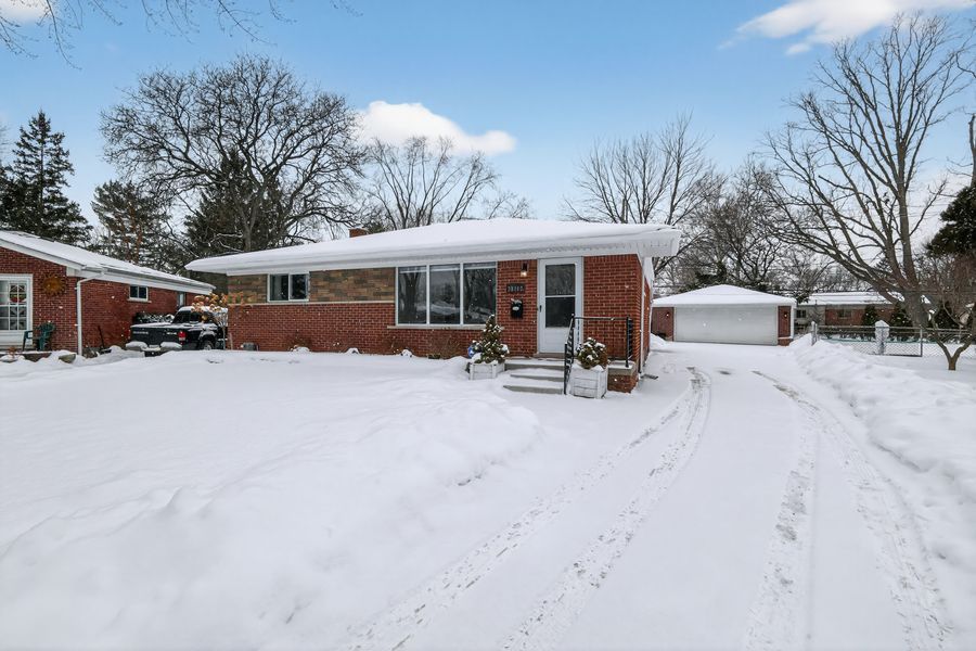 Brick ranch home with snow-covered yard, attached garage, and bare trees under a blue winter sky.