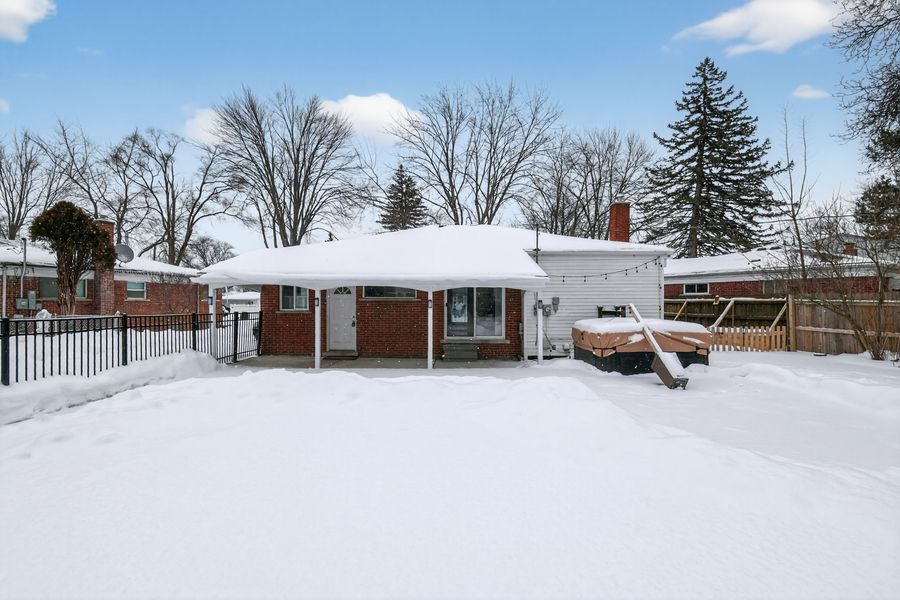 Snow-covered brick ranch home with white trim, large front yard, and mature trees under a bright blue winter sky.