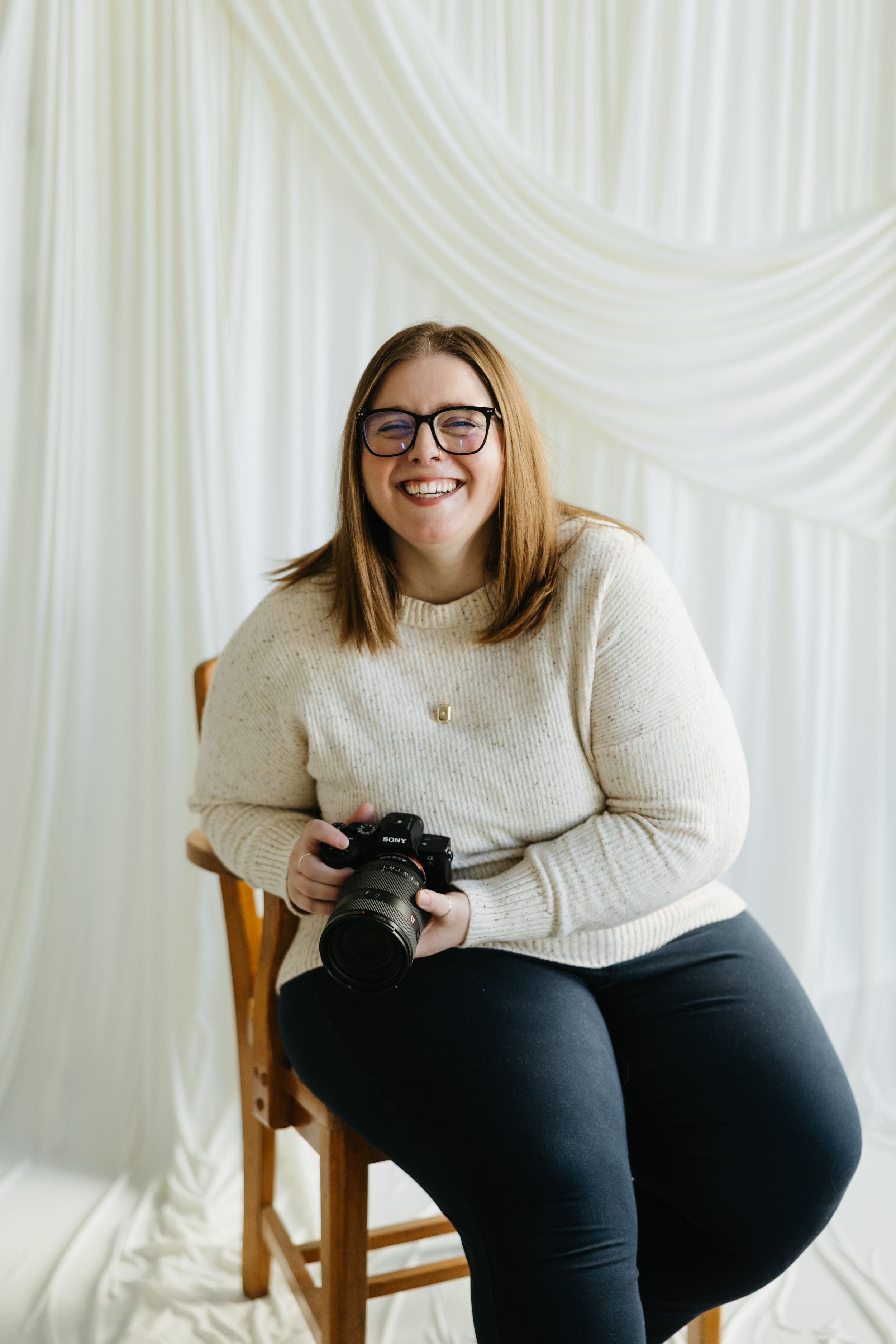 Smiling woman with glasses holding a camera, seated on a wooden chair against a white draped backdrop.