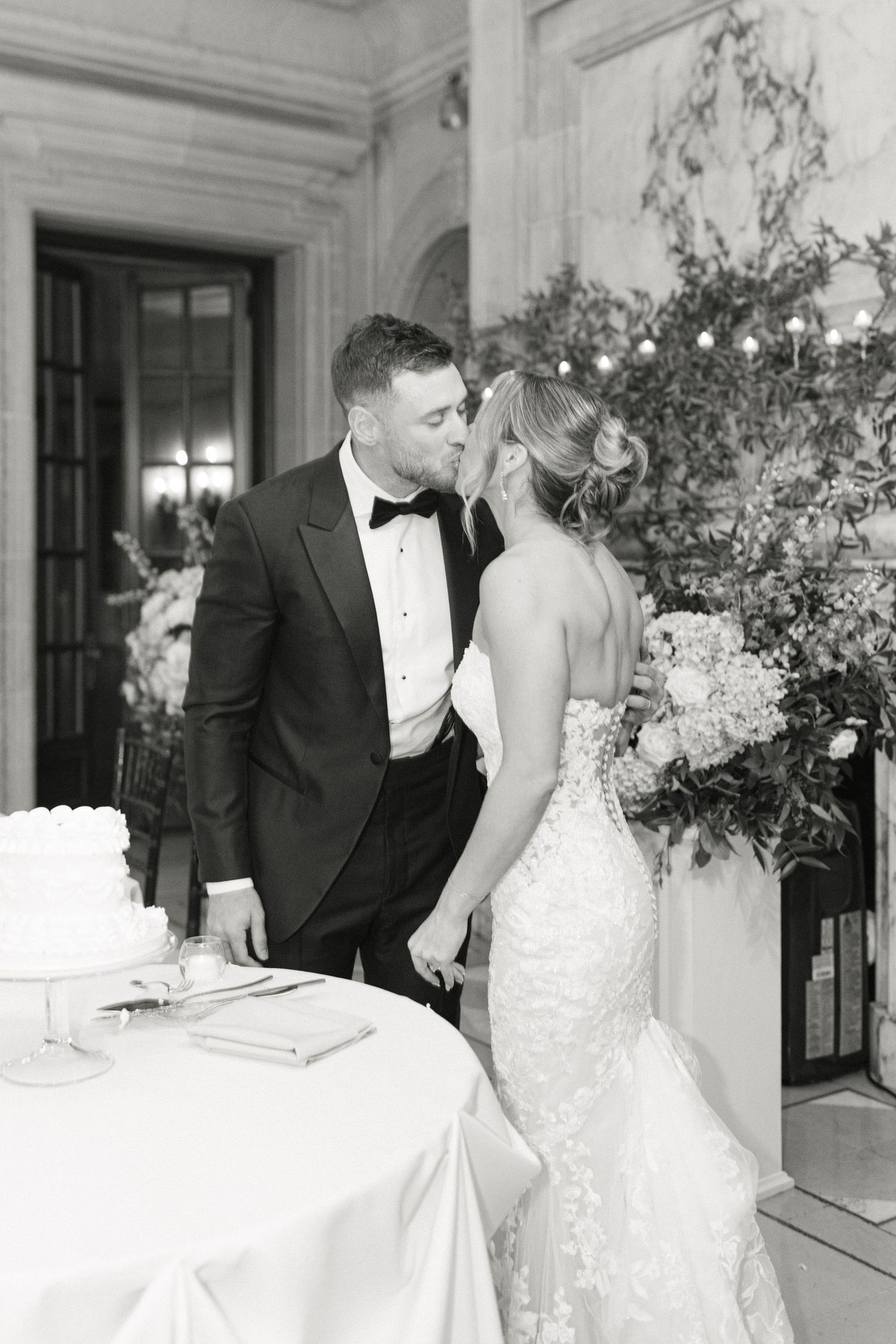 Black and white photo of bride and groom sharing a kiss beside their wedding cake at an elegant reception.