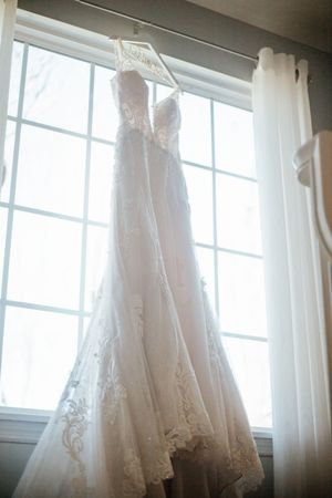 Elegant white lace A-line wedding dress hanging on a decorative hanger in front of a bright window with sheer curtains.