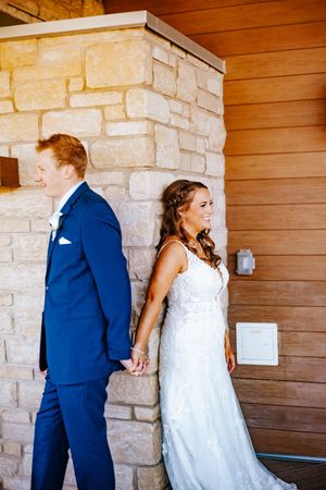 Bride in lace gown and groom in blue suit hold hands around a stone pillar at their wedding venue.