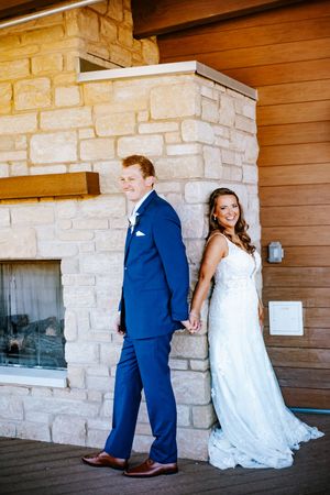 Bride in white lace gown and groom in blue suit hold hands, posing back-to-back by a stone fireplace.
