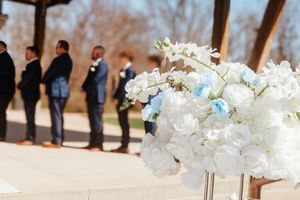 White and blue floral centerpiece at an outdoor wedding ceremony with groomsmen standing in the background.