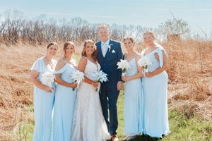 Bride and groom pose with four bridesmaids in light blue gowns holding white bouquets outdoors in a golden field.